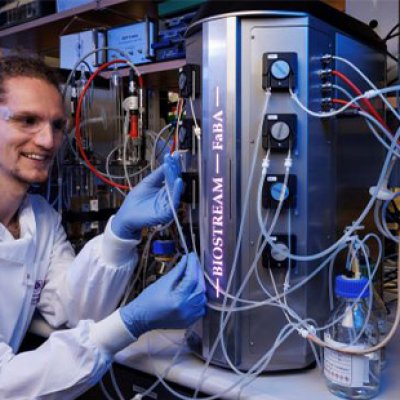 A man in a white coat and blue gloves sits in front of a silver cannister 